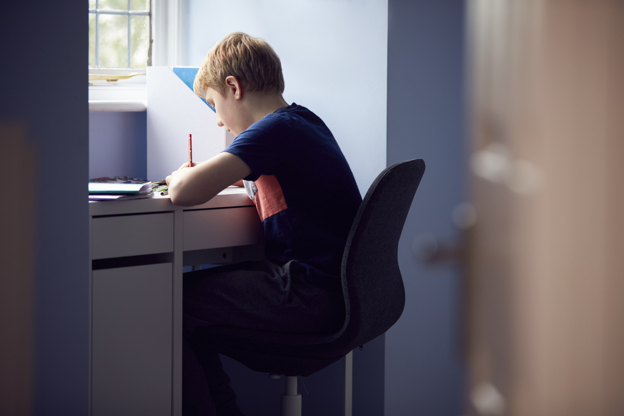 A caucasian boy working at a desk in front of a window.